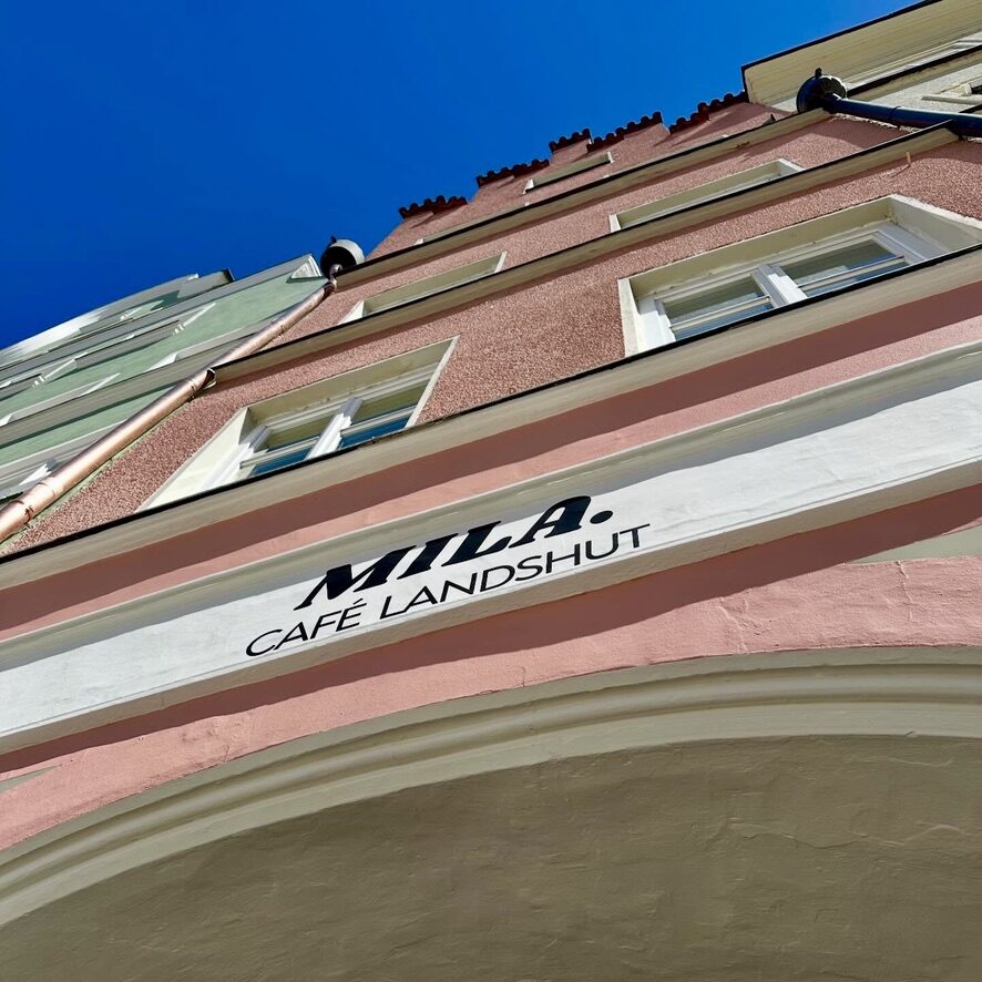 Die rosa Fassade des MILA. mit dem Schild „MILA. Café Landshut“, von unten gegen den blauen Himmel